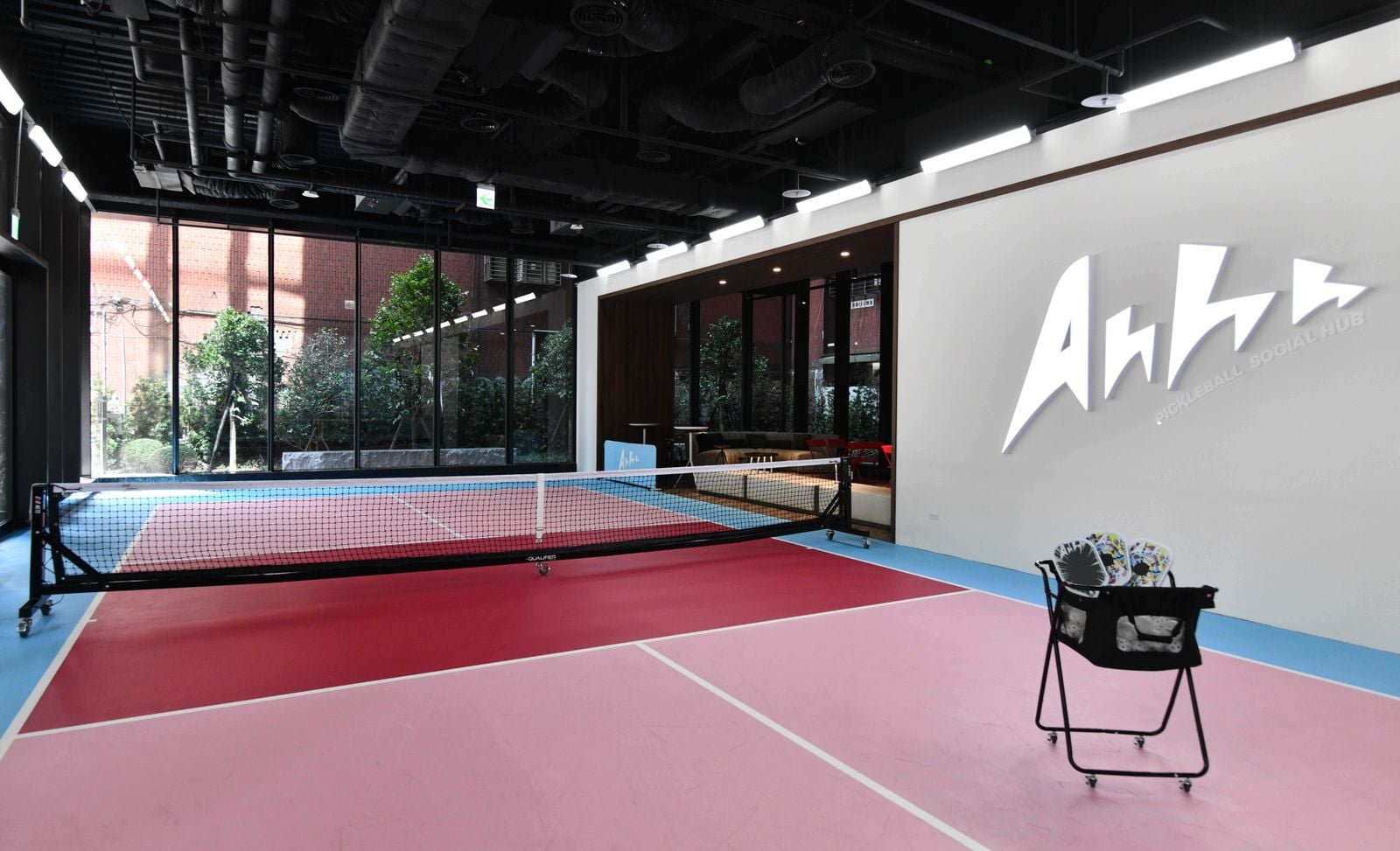 A brightly colored indoor pickleball court with a black net, a basket of balls, and an illuminated Ahhh logo on the wall.