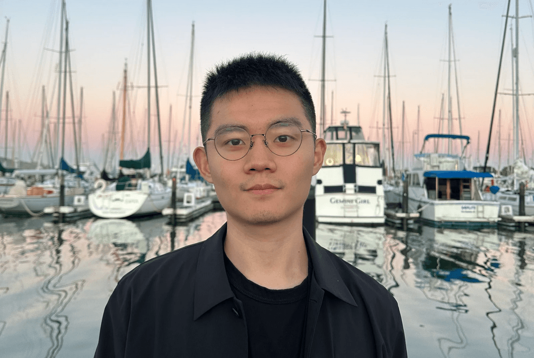 Cheng-Wei Hu stands in front of sailboats and their reflections at a marina around sunset.