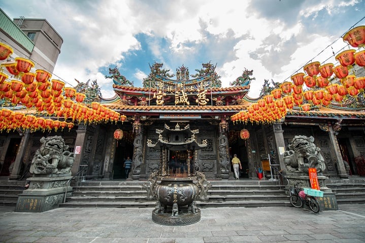 The front of Cixian Temple in Taipei, with rows of red and gold lanterns framing its carved roofline and a large incense burner centered before the steps.