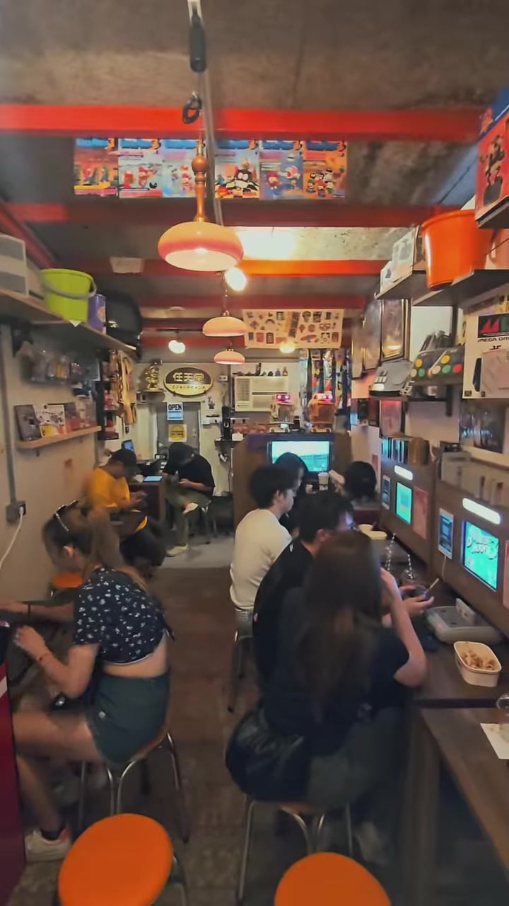 Visitors play retro games at counters lining a narrow Taipei cafe filled with old screens, game artwork, and orange pendant lamps.