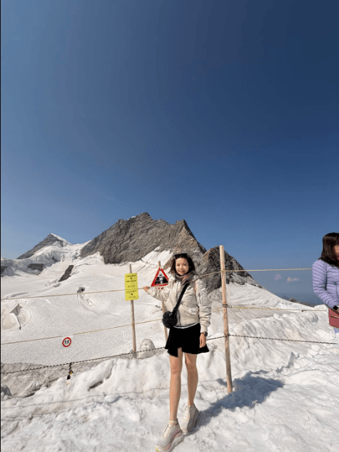 A woman in a silver jacket and black shorts stands on snow at Jungfrau in Switzerland, with rocky, snow-covered peaks and rope barriers behind her; another visitor is partly visible at right.