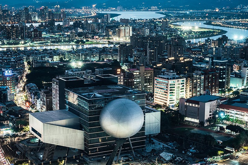 A nighttime aerial view shows Taipei Performing Arts Center’s silver spherical playhouse projecting from a dark central cube, with rectangular theater volumes and the illuminated city and river beyond.