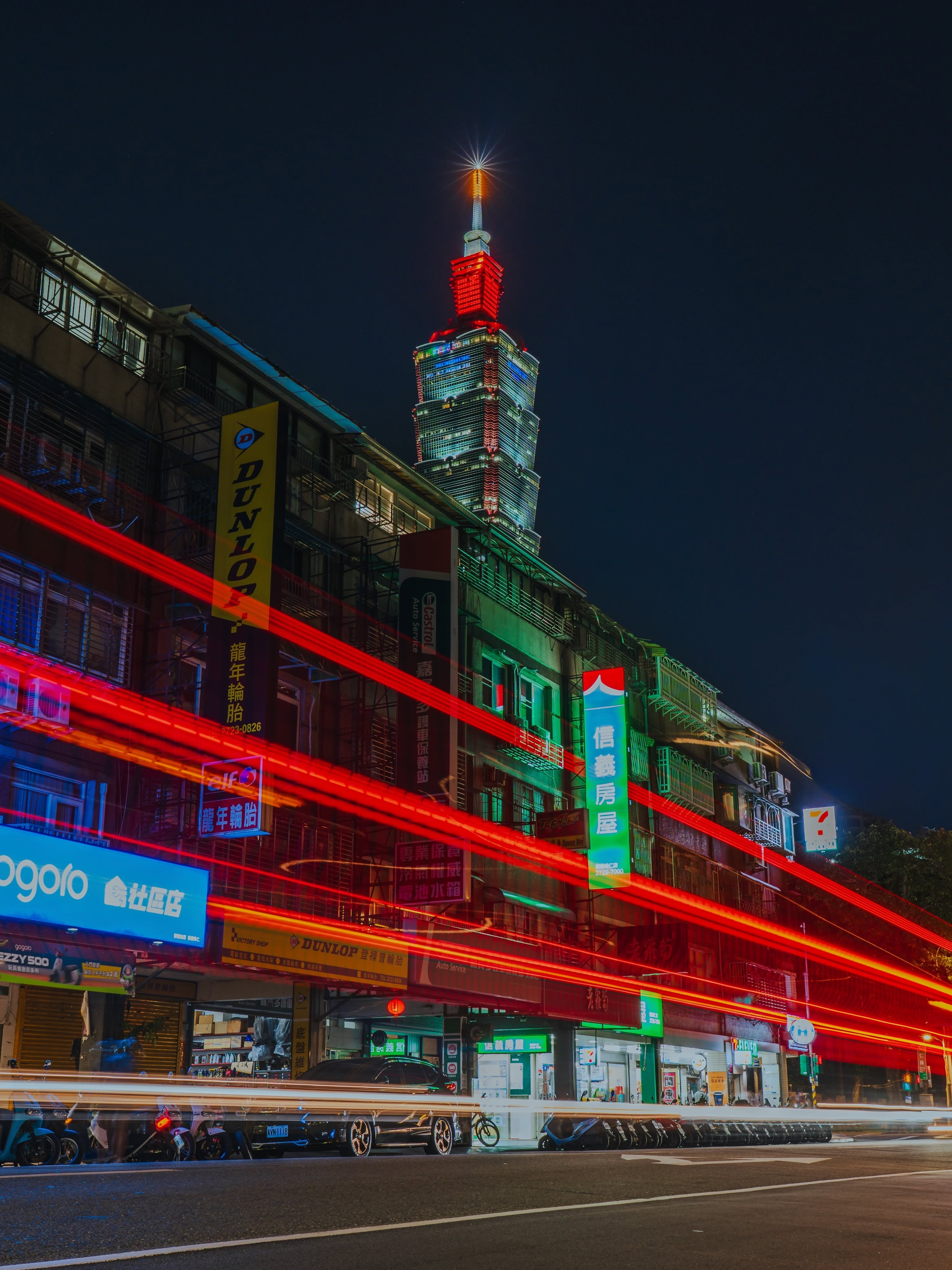 Taipei 101 rises above a night street filled with illuminated signs and streams of vehicle lights.