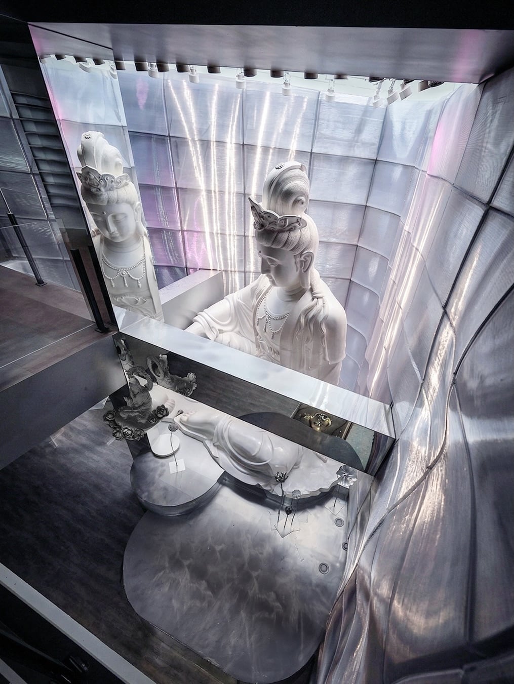 An overhead view of a tall white Guanyin statue spanning two levels inside reflective silver walls, with a reclining figure below.