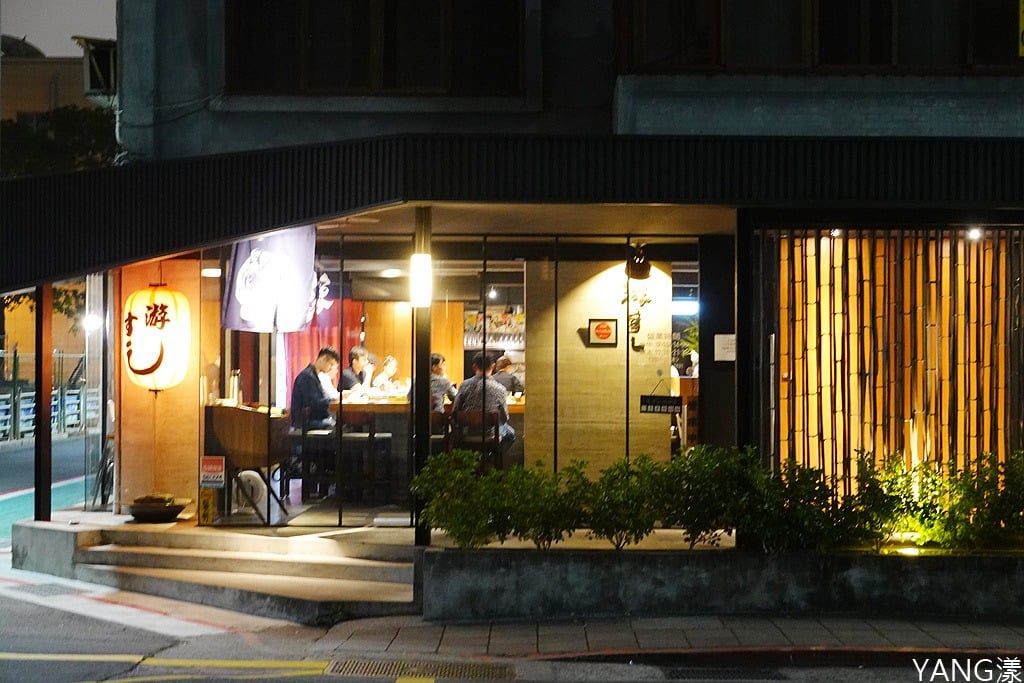 Diners sit behind the glass frontage of Yo Sushi Jinhua at night, beside an illuminated lantern bearing the restaurant's name.