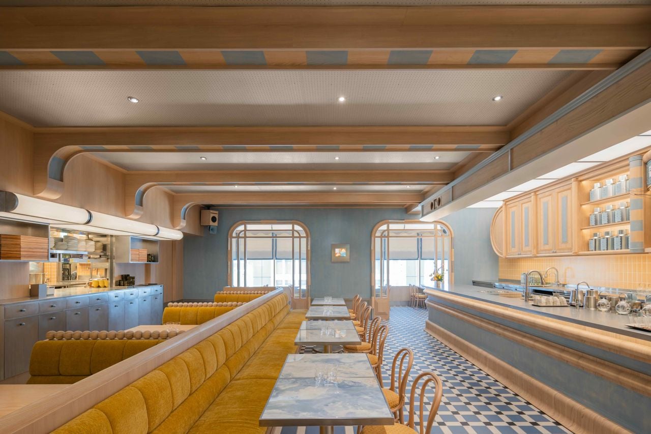 A pale blue and yellow dining room with arched openings, mustard banquettes, marble-top tables, and a long counter.