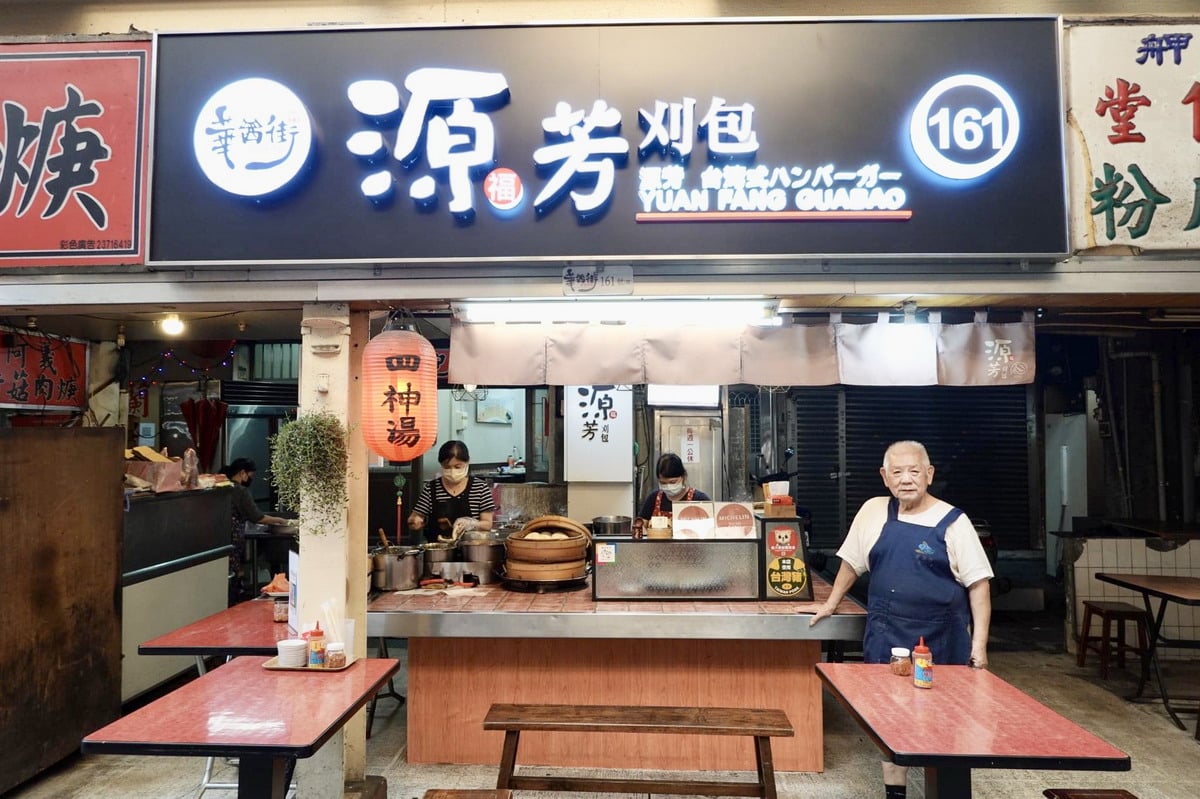 Staff work behind the Yuan Fang Guabao counter beneath an illuminated 源芳刈包 sign, while an older man stands beside the stall.