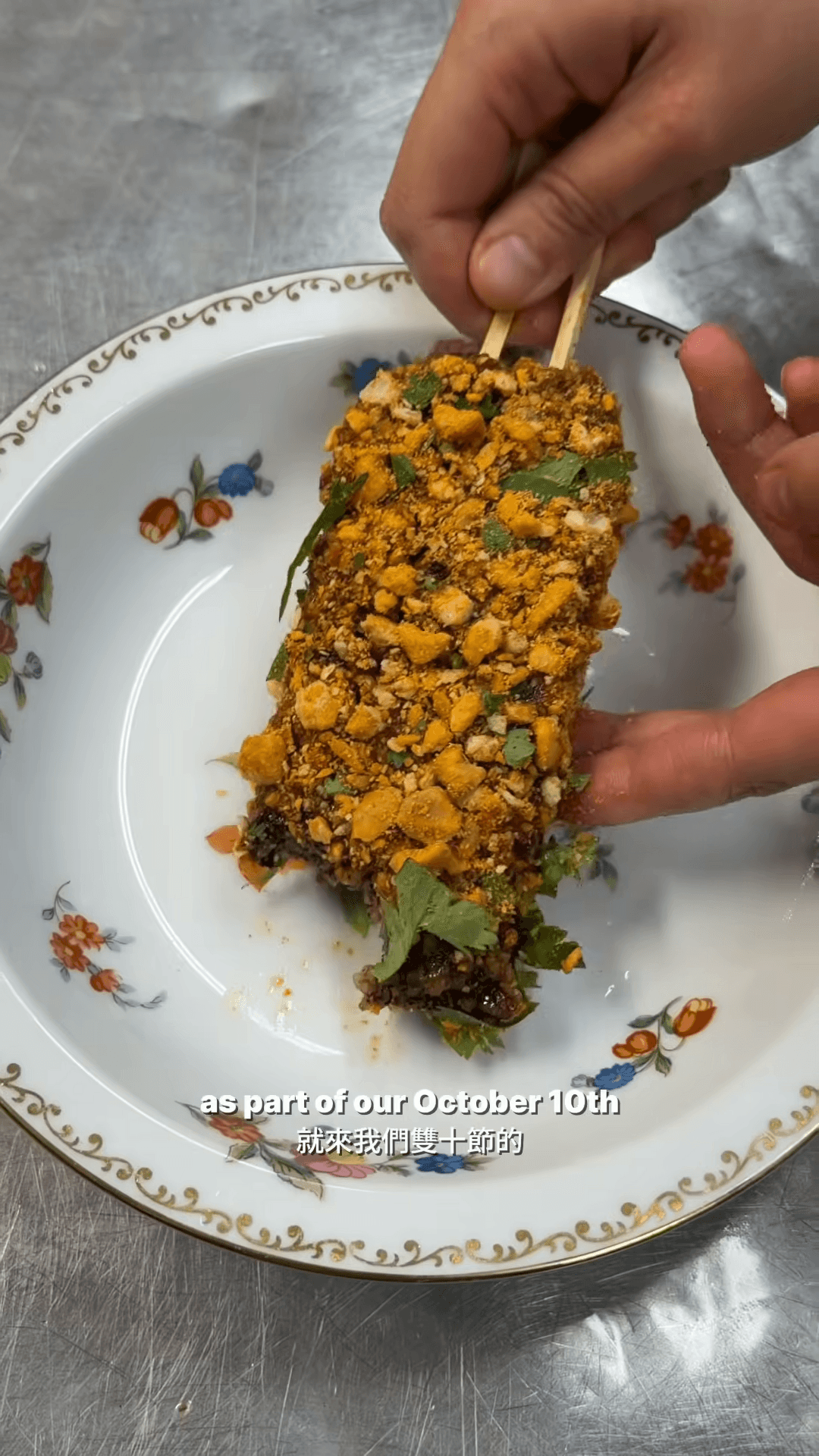 A rectangular piece of pig blood cake coated with crushed peanuts and cilantro rests on a floral plate as two wooden picks hold it upright.